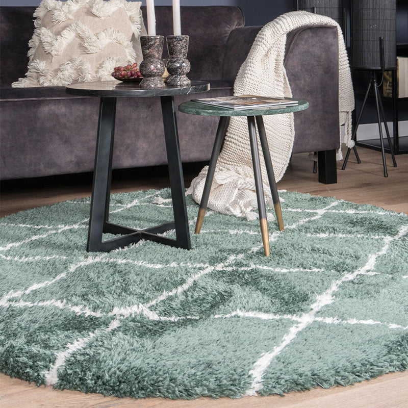 Groene vloerkleed met ruitpatroon, twee tafeltjes met decoratie, bruine bank en beige deken ernaast.